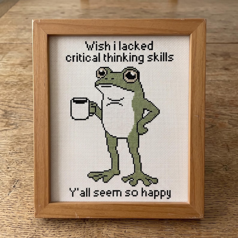 May include: A framed cross-stitch embroidery featuring a green frog holding a white mug. The text above reads "Wish I lacked critical thinking skills" and the text below reads "Y'all seem so happy". The piece is encased in a light wood frame.