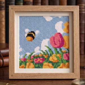 K&ouml;nnte beinhalten: Kreuzstich-Kunstwerk in einem Holzrahmen. Das Bild zeigt eine Biene, die in einem blauen Himmel mit wei&szlig;en Wolken &uuml;ber einem Feld bunter Blumen fliegt. Die Blumen umfassen rosa, gelbe und orangefarbene Bl&uuml;ten.
