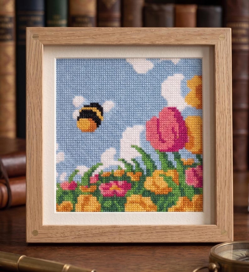 K&ouml;nnte beinhalten: Kreuzstich-Kunstwerk in einem Holzrahmen. Das Bild zeigt eine Biene, die in einem blauen Himmel mit wei&szlig;en Wolken &uuml;ber einem Feld bunter Blumen fliegt. Die Blumen umfassen rosa, gelbe und orangefarbene Bl&uuml;ten.