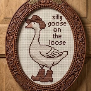 Op de afbeelding: Kruissteekkunstwerk van een gans met een cowboyhoed en laarzen, met de tekst "silly goose on the loose". Het kunstwerk bevindt zich in een ovale, sierlijke, bruine houten lijst, met een decoratieve metalen hanger.