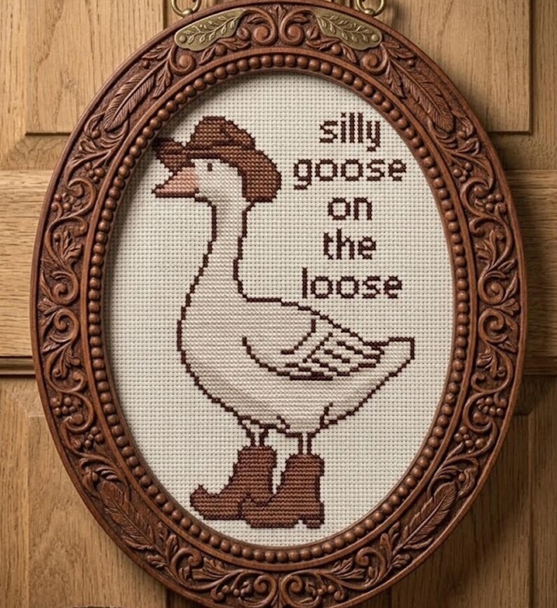 Op de afbeelding: Kruissteekkunstwerk van een gans met een cowboyhoed en laarzen, met de tekst "silly goose on the loose". Het kunstwerk bevindt zich in een ovale, sierlijke, bruine houten lijst, met een decoratieve metalen hanger.