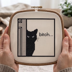 Puede incluir: Un bastidor de bordado cuadrado de madera que contiene un dise&ntilde;o de punto de cruz sobre tela blanca. El dise&ntilde;o muestra un gato negro asom&aacute;ndose detr&aacute;s de un marco de puerta gris con el texto "bitch..." bordado en hilo negro a la derecha.