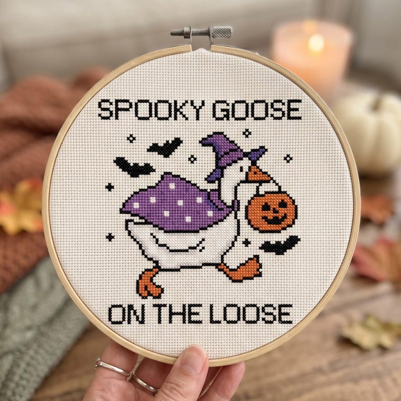 May include: A circular wooden embroidery hoop containing a cross-stitch design of a white goose wearing a purple polka-dot cape and a purple witch hat. The goose carries an orange jack-o'-lantern bucket. Text reads SPOOKY GOOSE ON THE LOOSE.