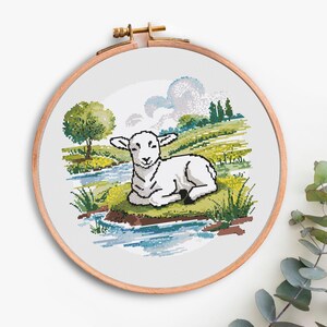 May include: A cross-stitch embroidery hoop featuring a pastoral scene. A white lamb rests on a grassy bank beside a stream. The background includes a tree, rolling hills, and a cloudy sky. The wooden hoop is round.