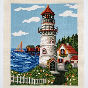 Peut inclure: Une broderie au point de croix repr&eacute;sentant un phare blanc au toit rouge, une petite maison au toit rouge, une mer bleue avec un voilier et une cl&ocirc;ture blanche entour&eacute;e de feuillage vert et de petites fleurs orange et blanches sous un ciel bleu.