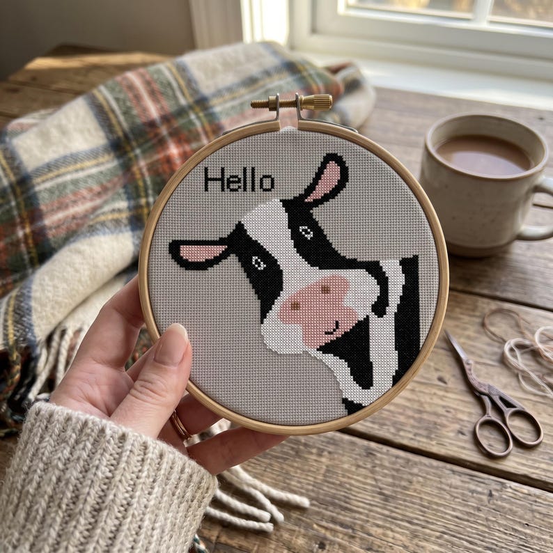 May include: A circular wooden embroidery hoop containing a cross-stitch pattern of a black and white cow on a light gray fabric background. The cow has a pink nose and ears, and the word Hello is stitched in black thread above the cow's head.