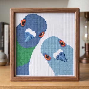 May include: A square cross-stitch embroidery featuring two pigeons in a wooden frame. The left pigeon is dark blue with a green neck patch, while the right pigeon is light blue. Both birds have orange eyes and white markings above their beaks.