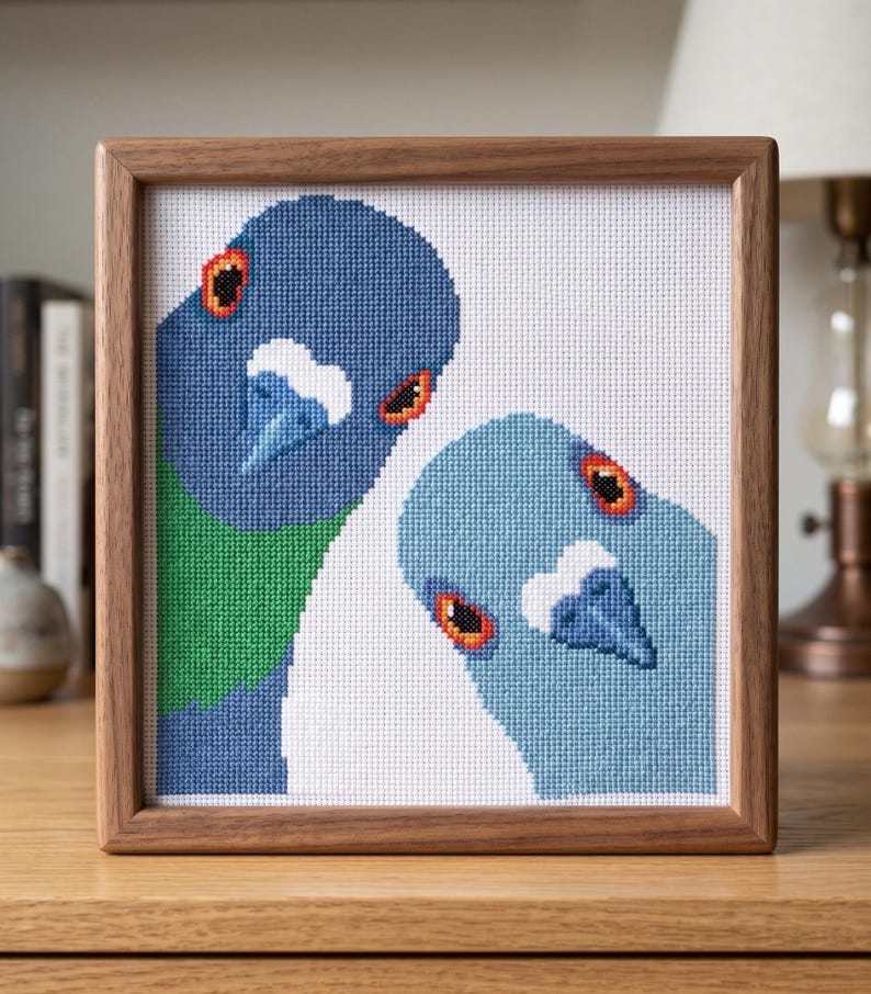 May include: A square cross-stitch embroidery featuring two pigeons in a wooden frame. The left pigeon is dark blue with a green neck patch, while the right pigeon is light blue. Both birds have orange eyes and white markings above their beaks.