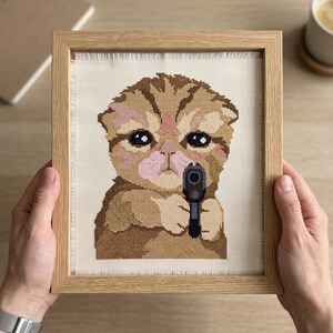Puede incluir: Un bordado en punto de cruz enmarcado que muestra un gatito atigrado marr&oacute;n con grandes ojos oscuros y nariz rosa, sosteniendo una peque&ntilde;a pistola negra. La obra est&aacute; bordada sobre tela Aida color hueso con bordes deshilachados en un marco de madera clara.