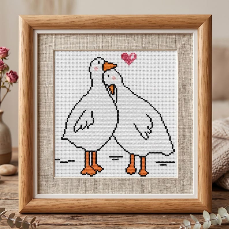 Puede incluir: Obra de arte de punto de cruz de dos gansos blancos abraz&aacute;ndose, con picos y patas naranjas, y un coraz&oacute;n rosa encima. La pieza enmarcada tiene un marco de madera marr&oacute;n claro y un paspart&uacute; de tela neutra.