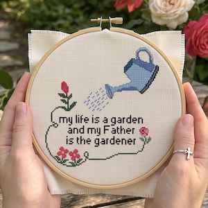 Op de afbeelding: Een ronde houten borduurring met witte Aida-stof en een kruissteekontwerp. Het ontwerp toont een lichtblauwe gieter, roze bloemen en de zwarte tekst: my life is a garden and my Father is the gardener.