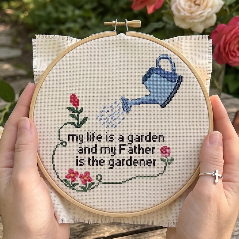 Op de afbeelding: Een ronde houten borduurring met witte Aida-stof en een kruissteekontwerp. Het ontwerp toont een lichtblauwe gieter, roze bloemen en de zwarte tekst: my life is a garden and my Father is the gardener.