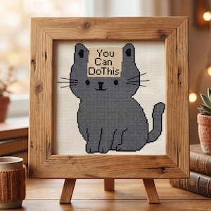 May include: Cross-stitch art of a grey cat with the text "You Can Do This" on a small sign above its head. The artwork is displayed in a rustic wooden frame on a small easel.