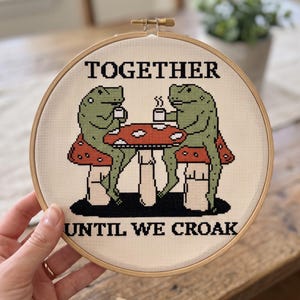 Pu&ograve; includere: Un telaio da ricamo circolare in legno contenente un motivo a punto croce su tessuto color crema. Il design presenta due rane verdi sedute a un tavolo a forma di fungo rosso che bevono da tazze bianche, con il testo TOGETHER UNTIL WE CROAK.