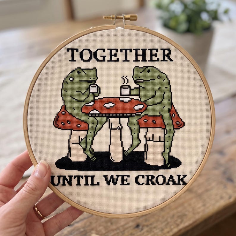 Pu&ograve; includere: Un telaio da ricamo circolare in legno contenente un motivo a punto croce su tessuto color crema. Il design presenta due rane verdi sedute a un tavolo a forma di fungo rosso che bevono da tazze bianche, con il testo TOGETHER UNTIL WE CROAK.