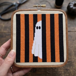 May include: A square wooden embroidery hoop containing a cross-stitch pattern of a white ghost centred against a background of alternating vertical black and orange stripes on a cream-coloured fabric base, secured with a metal tension screw.