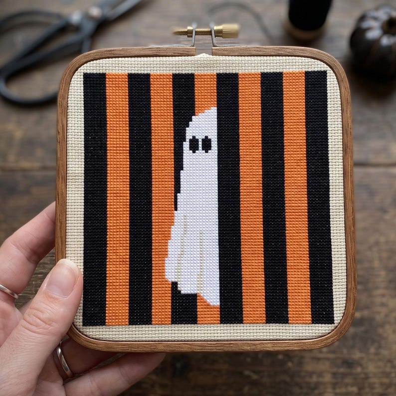 May include: A square wooden embroidery hoop containing a cross-stitch pattern of a white ghost centred against a background of alternating vertical black and orange stripes on a cream-coloured fabric base, secured with a metal tension screw.