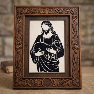 Pode incluir: Uma imagem bordada emoldurada que retrata uma figura religiosa segurando um cordeiro. A obra de arte em preto e branco est&aacute; emoldurada em uma moldura de madeira marrom escura com detalhes esculpidos de uvas e trigo. A moldura tem formato retangular.