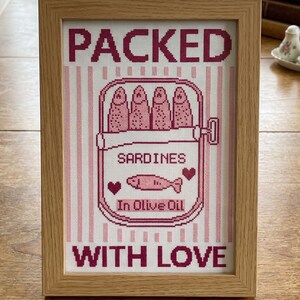 Pu&ograve; includere: Un'opera d'arte a punto croce incorniciata presenta una scatola di sardine con le parole "PACKED WITH LOVE". Il design include sardine rosa, cuori e il testo "SARDINES In Olive Oil". La cornice &egrave; in legno chiaro.