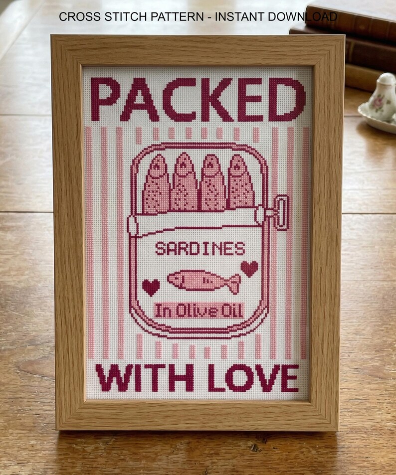 Pu&ograve; includere: Un'opera d'arte a punto croce incorniciata presenta una scatola di sardine con le parole "PACKED WITH LOVE". Il design include sardine rosa, cuori e il testo "SARDINES In Olive Oil". La cornice &egrave; in legno chiaro.