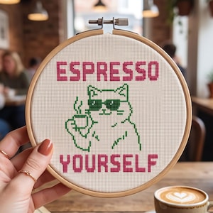 Puede incluir: Un bastidor de bordado circular de madera que contiene una tela de color blanco roto con un dise&ntilde;o de punto de cruz. El dise&ntilde;o muestra un gato verde con gafas de sol sosteniendo una taza de caf&eacute;, con el texto rosa ESPRESSO YOURSELF.