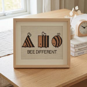 May include: A framed cross-stitch embroidery featuring three geometric bee shapes in black and tan, including a triangle, a square, and a circle. Below the bees, the text reads "BEE DIFFERENT" in black capital letters on a light fabric background.