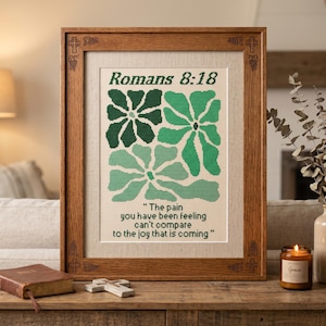 May include: A framed cross-stitch artwork in a wooden frame. The artwork displays green floral designs and the text "Romans 8:18" and a quote. A bible, a cross, and a candle are on the wooden surface in front of the artwork.