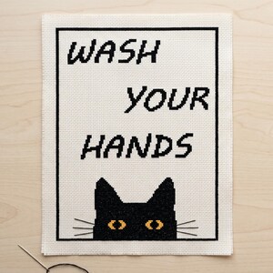 K&ouml;nnte beinhalten: Eine Kreuzstichstickerei auf wei&szlig;em Aida-Stoff mit einem schwarzen rechteckigen Rahmen. Der Text WASH YOUR HANDS ist in schwarzen Blockbuchstaben gestickt. Unter dem Text befindet sich die Silhouette einer schwarzen Katze mit gelben Augen.