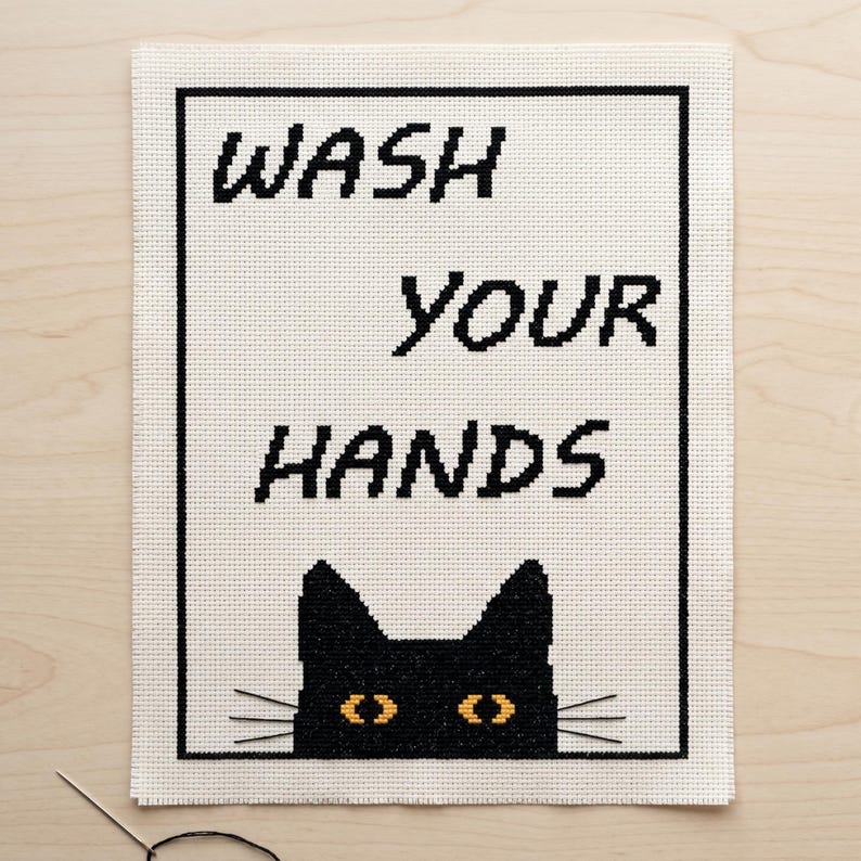 K&ouml;nnte beinhalten: Eine Kreuzstichstickerei auf wei&szlig;em Aida-Stoff mit einem schwarzen rechteckigen Rahmen. Der Text WASH YOUR HANDS ist in schwarzen Blockbuchstaben gestickt. Unter dem Text befindet sich die Silhouette einer schwarzen Katze mit gelben Augen.