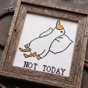 May include: Cross-stitch artwork in a rustic wooden frame. The design features a white cartoon character with a yellow hat and feet, resting on a pillow. The text "NOT TODAY" is stitched in black below the character.