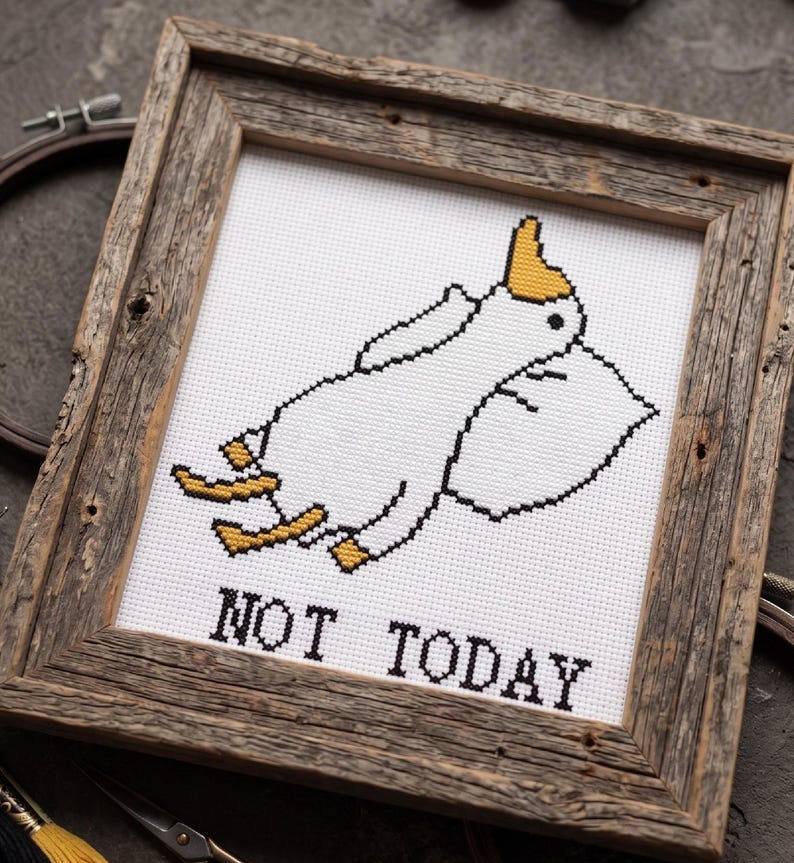 May include: Cross-stitch artwork in a rustic wooden frame. The design features a white cartoon character with a yellow hat and feet, resting on a pillow. The text "NOT TODAY" is stitched in black below the character.
