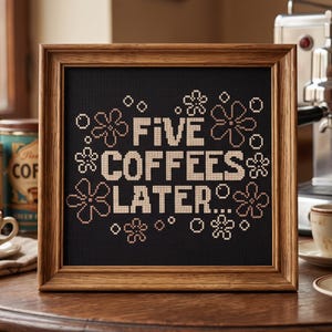 May include: A square cross-stitch embroidery framed in wood, featuring the text "FIVE COFFEES LATER..." in beige thread against a black fabric background, surrounded by stitched beige flower and bubble motifs in a decorative arrangement.