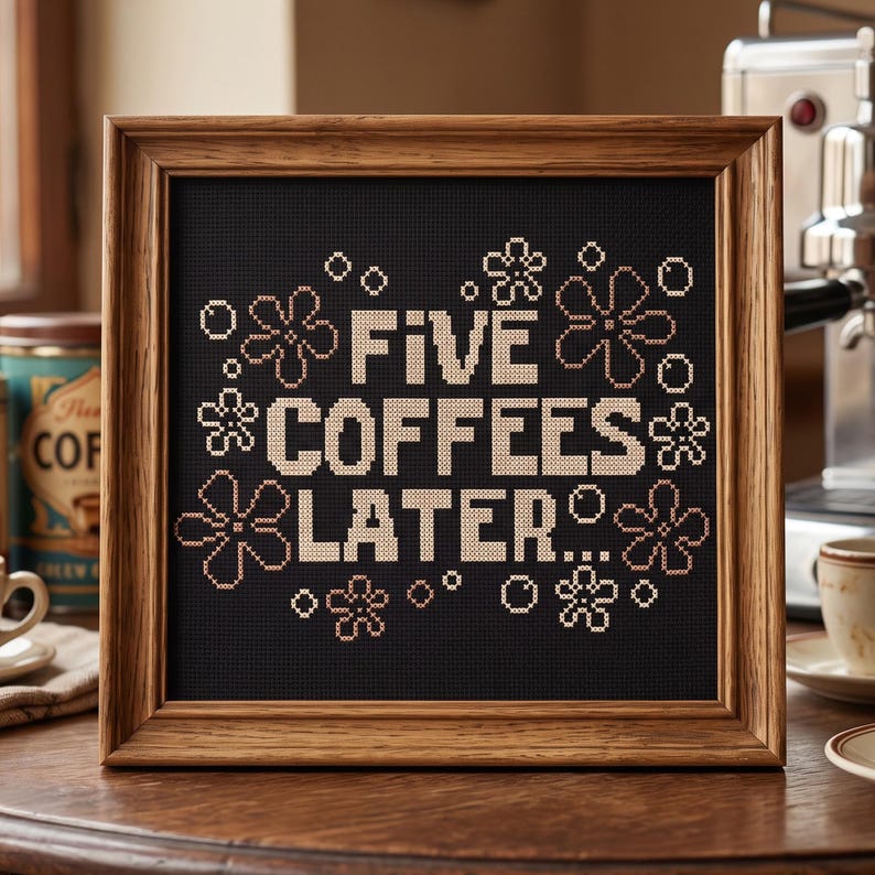 May include: A square cross-stitch embroidery framed in wood, featuring the text "FIVE COFFEES LATER..." in beige thread against a black fabric background, surrounded by stitched beige flower and bubble motifs in a decorative arrangement.