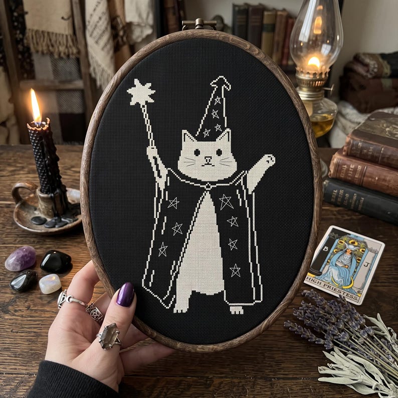 May include: An oval embroidery hoop containing a black fabric piece with a white cross-stitch design of a cat dressed as a wizard. The cat wears a star-patterned robe and hat, holding a star-tipped wand. The hoop has a dark brown wooden frame.