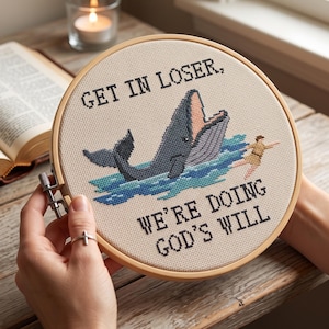 May include: A circular wooden embroidery hoop containing a cross-stitch design on cream fabric. The design features a dark gray whale with an open mouth, a small figure in the blue water, and the black text: GET IN LOSER, WE'RE DOING GOD'S WILL.