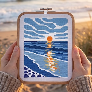 Pu&ograve; includere: Un telaio da ricamo rettangolare in legno contenente un disegno a punto croce di un tramonto sull'oceano. La scena presenta un sole arancione brillante, onde blu scuro e un cielo azzurro con nuvole, con il riflesso del sole sull'acqua.