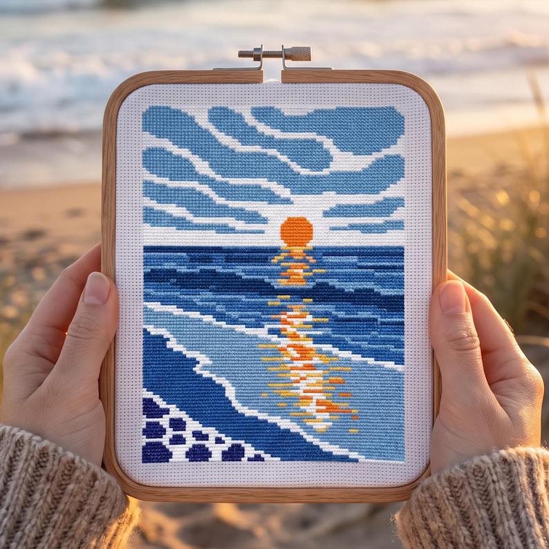 Pu&ograve; includere: Un telaio da ricamo rettangolare in legno contenente un disegno a punto croce di un tramonto sull'oceano. La scena presenta un sole arancione brillante, onde blu scuro e un cielo azzurro con nuvole, con il riflesso del sole sull'acqua.
