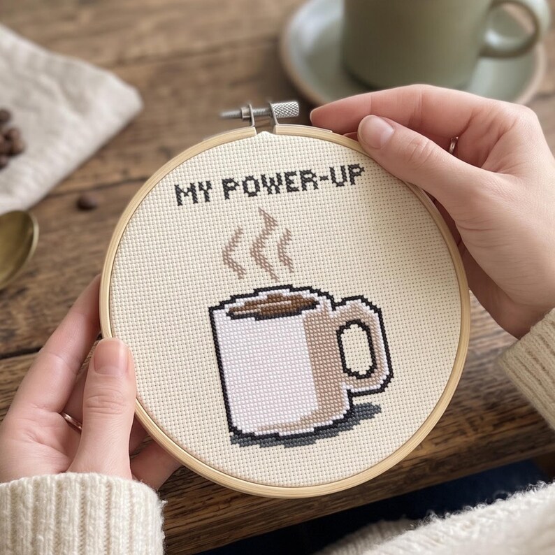 Puede incluir: Un bastidor de bordado circular de madera que contiene un dise&ntilde;o de punto de cruz sobre tela de color crema. El dise&ntilde;o presenta una taza de caf&eacute; blanca con l&iacute;quido marr&oacute;n y vapor elev&aacute;ndose, junto al texto negro MY POWER-UP sobre la taza.