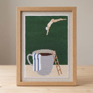 Puede incluir: Un bordado en punto de cruz enmarcado con fondo verde oscuro que muestra una figura saltando hacia una taza de caf&eacute; grande de color gris claro. La taza tiene una toalla a rayas blancas y azules y una escalera marr&oacute;n apoyada en el lateral.