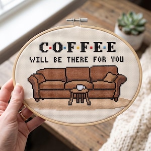 May include: An oval wooden embroidery hoop containing a cross-stitch design on cream fabric. The design features a brown sofa, a small table with a white cup, and the black text COFFEE WILL BE THERE FOR YOU with colorful dots between letters.