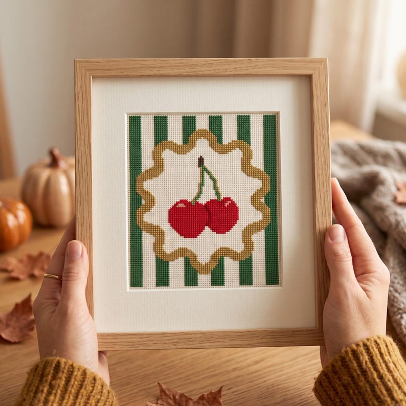 May include: A framed cross-stitch embroidery featuring two bright red cherries with green stems centred on a white background with vertical dark green stripes, enclosed within a scalloped gold border, all set in a light-toned wooden frame.