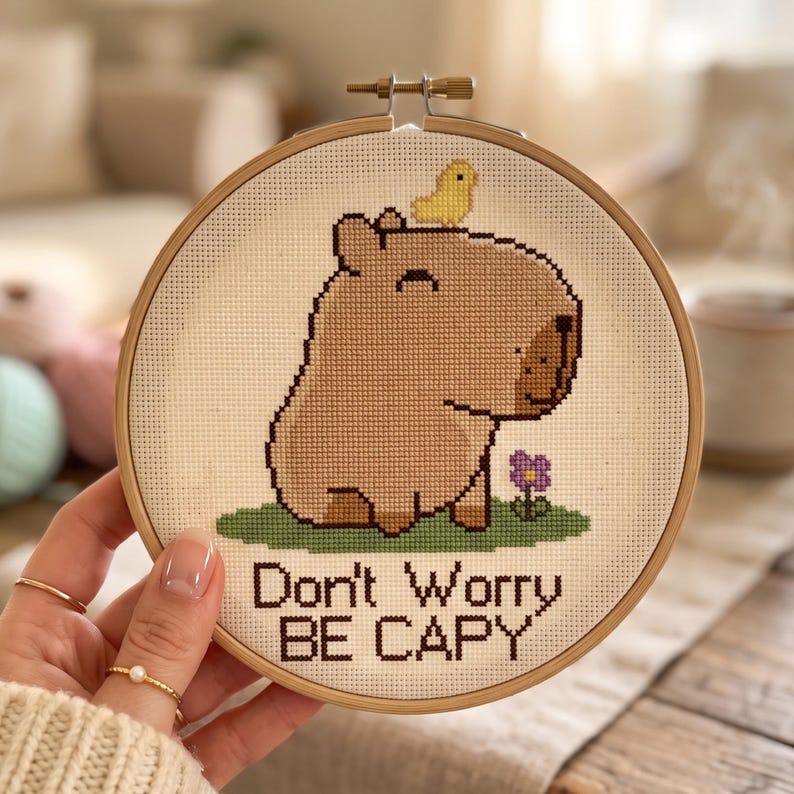 May include: A circular wooden embroidery hoop containing a cross-stitch design of a light brown capybara with a small yellow bird on its head, standing on green grass next to a purple flower, with the text Don't Worry BE CAPY stitched below.