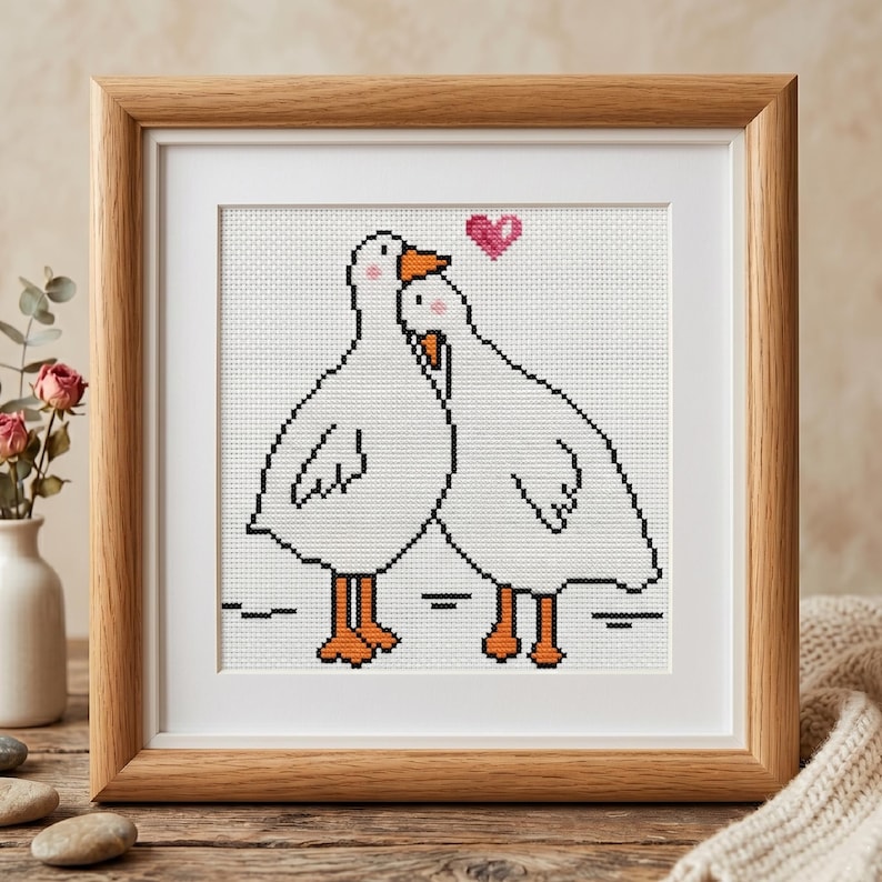 Puede incluir: Una obra de arte en punto de cruz enmarcada que presenta dos gansos blancos abraz&aacute;ndose, con picos y patas naranjas. Un coraz&oacute;n rosa flota sobre ellos. La obra de arte est&aacute; en un marco de madera.