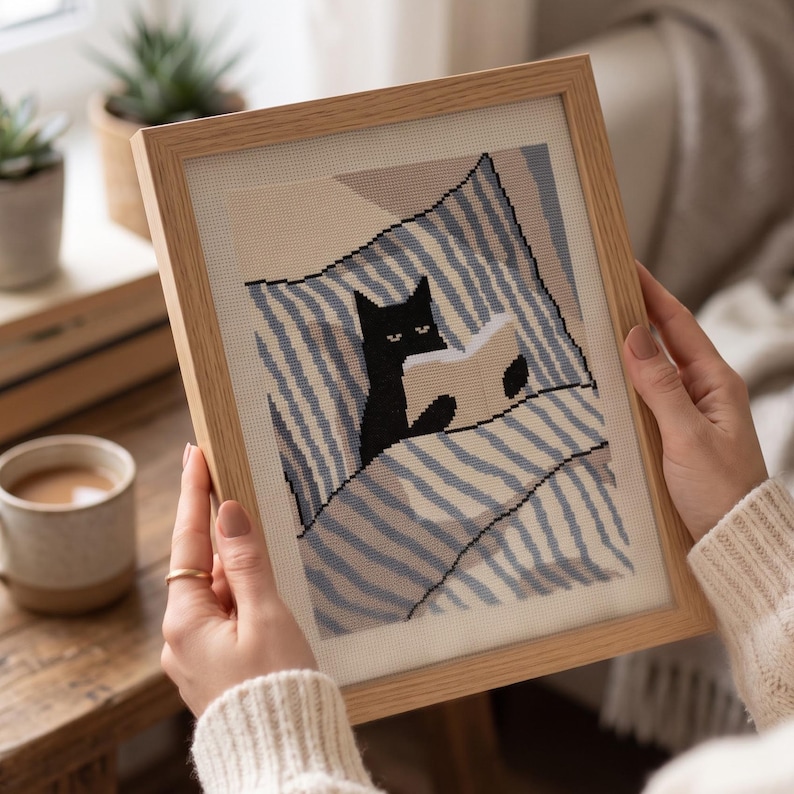 Op de afbeelding: Een ingelijst kruissteek borduurwerk met een zwarte kat die in bed ligt met blauw-wit gestreept beddengoed. De kat houdt een open boek vast, waarbij de kop en pootjes boven de dekens uitsteken. Het kunstwerk zit in een lichthouten lijst.