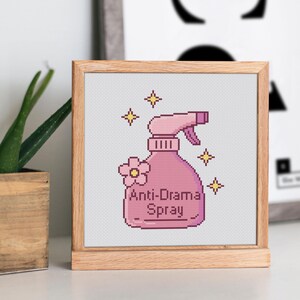 Peut inclure: Une broderie au point de croix encadr&eacute;e repr&eacute;sentant un flacon pulv&eacute;risateur rose avec le texte "Anti-Drama Spray". La bouteille est orn&eacute;e d'une fleur et d'&eacute;clairs. Le cadre est en bois clair.