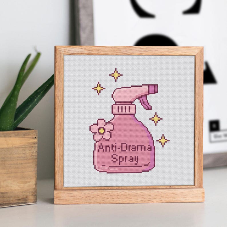 Peut inclure: Une broderie au point de croix encadr&eacute;e repr&eacute;sentant un flacon pulv&eacute;risateur rose avec le texte "Anti-Drama Spray". La bouteille est orn&eacute;e d'une fleur et d'&eacute;clairs. Le cadre est en bois clair.