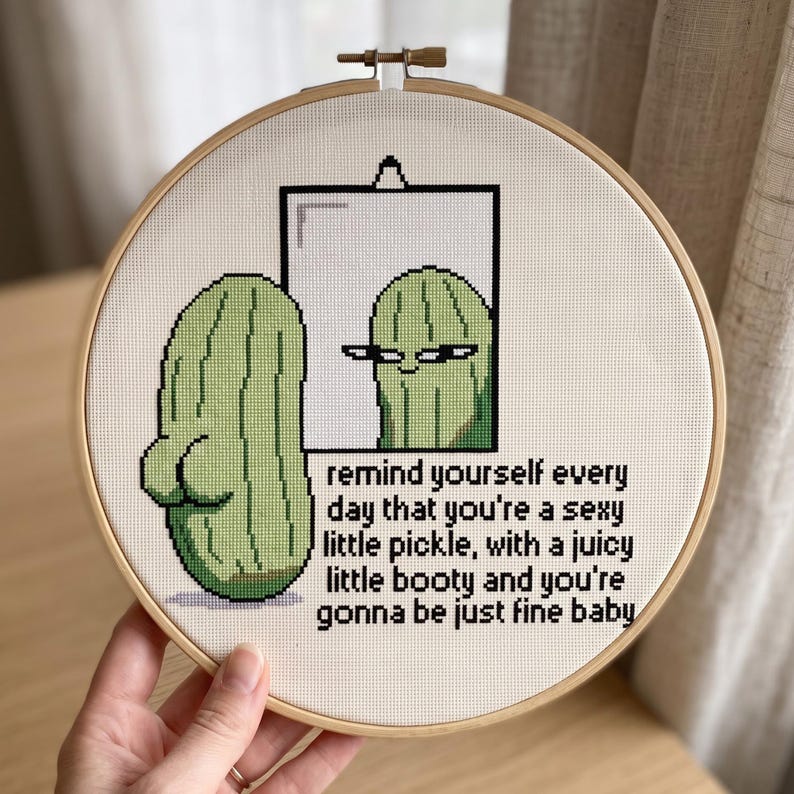 May include: A circular wooden embroidery hoop containing a cross-stitch design of a green pickle looking into a mirror. The pickle has a prominent backside. The text reads: remind yourself every day that you're a sexy little pickle, with a juicy little booty and you're gonna be just fine baby.