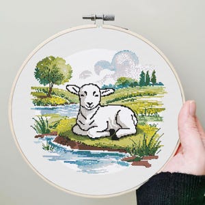 May include: A cross-stitch embroidery hoop featuring a pastoral scene. A white lamb rests on a grassy knoll near a stream, with trees and clouds in the background. The embroidery is set in a wooden hoop.