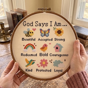 K&ouml;nnte beinhalten: Ein runder Holzstickrahmen mit einem Kreuzstich-Muster auf cremefarbenem Stoff. Der Text lautet God Says I Am... gefolgt von neun Reihen mit Symbolen und W&ouml;rtern: Beautiful, Accepted, Strong, Redeemed, Bold, Courageous, Kind, Protected, Loyal.