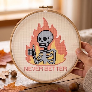 May include: A circular wooden embroidery hoop containing a cross-stitch design of a light blue skeleton giving a thumbs-up gesture, surrounded by orange and yellow flames, with the text NEVER BETTER stitched in red letters below the skeleton.